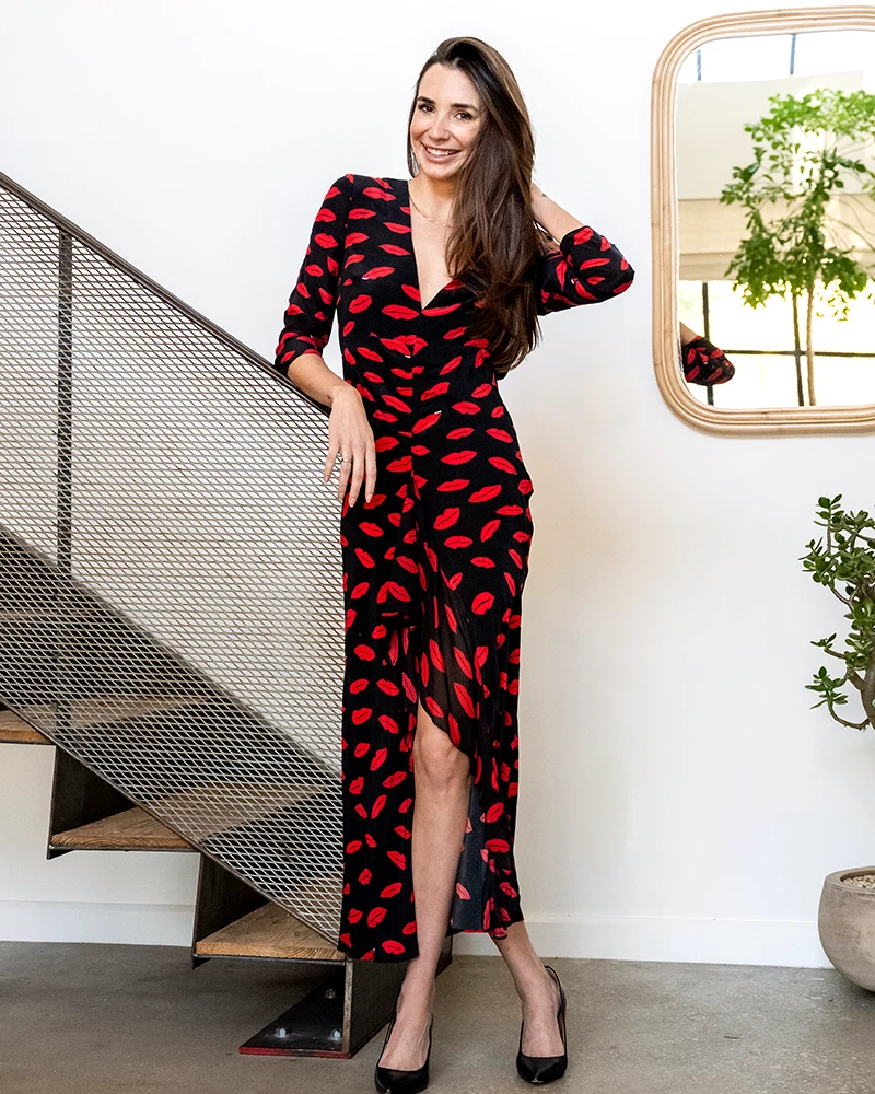 Photo of online dating coach Blaine Anderson, in a black dress with a red print, leaning against a staircase