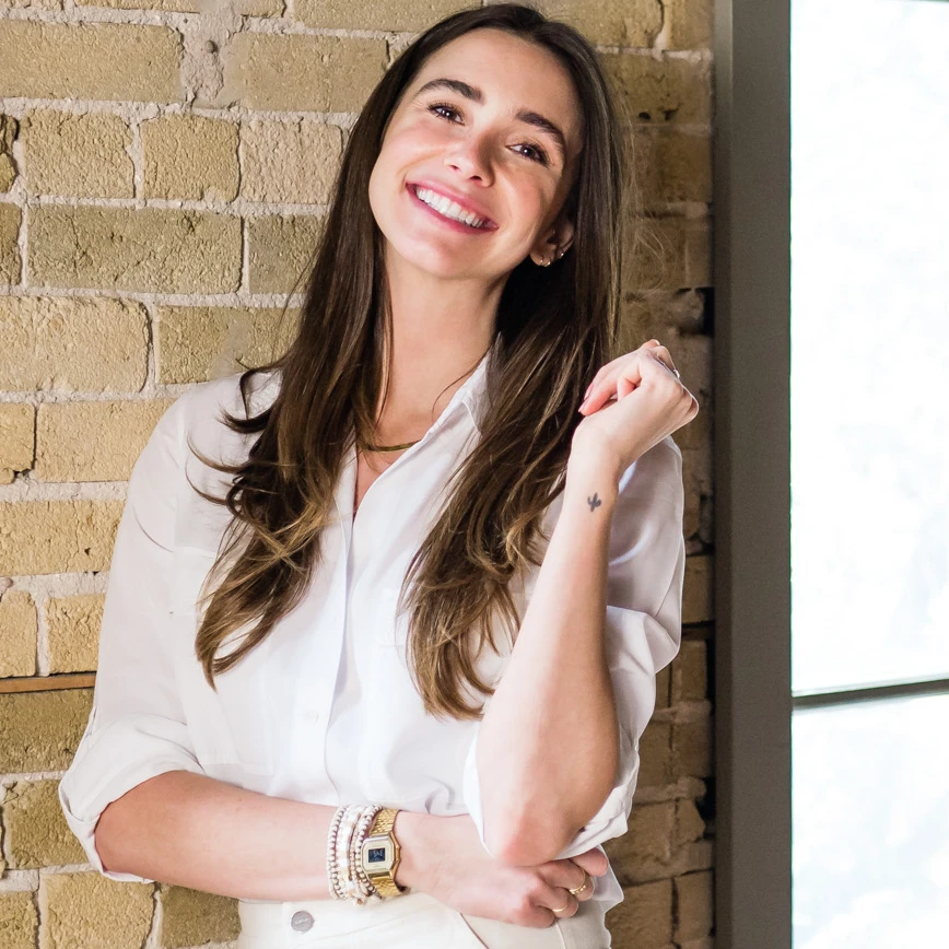 Photo of Blaine in a white outfit in a loft-like building, standing, smiling, head tilted.